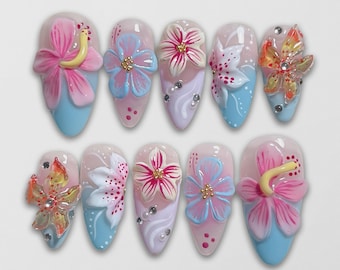 May include: A set of ten artificial nails, each adorned with a unique 3D floral design. The nails feature a variety of colors, including pink, blue, white, and yellow, with accents of rhinestones and glitter. The nail shapes are oval.