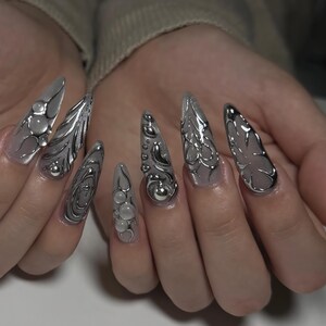 May include: A set of long, pointed fingernails with a silver metallic design. The nails feature various 3D embellishments, including spheres and intricate, flowing patterns. The base of the nails is a translucent, light color.