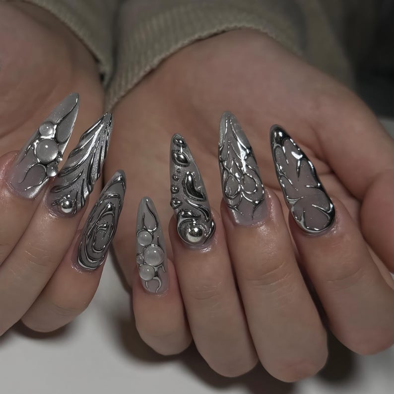 May include: A set of long, pointed fingernails with a silver metallic design. The nails feature various 3D embellishments, including spheres and intricate, flowing patterns. The base of the nails is a translucent, light color.