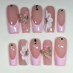 Puede incluir: Un conjunto de diez uñas artificiales con varios diseños. Las uñas presentan una base rosa claro con una punta francesa rosa más clara. Algunas uñas tienen un adorno de mariposa o flor, y todas tienen una pequeña gema.