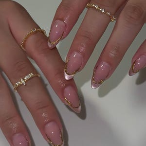 May include: Almond-shaped nails featuring a sheer nude base with a metallic gold French tip outline and a subtle cat-eye shimmer effect. The fingers are adorned with three thin gold rings, including one with a small clear stone setting.