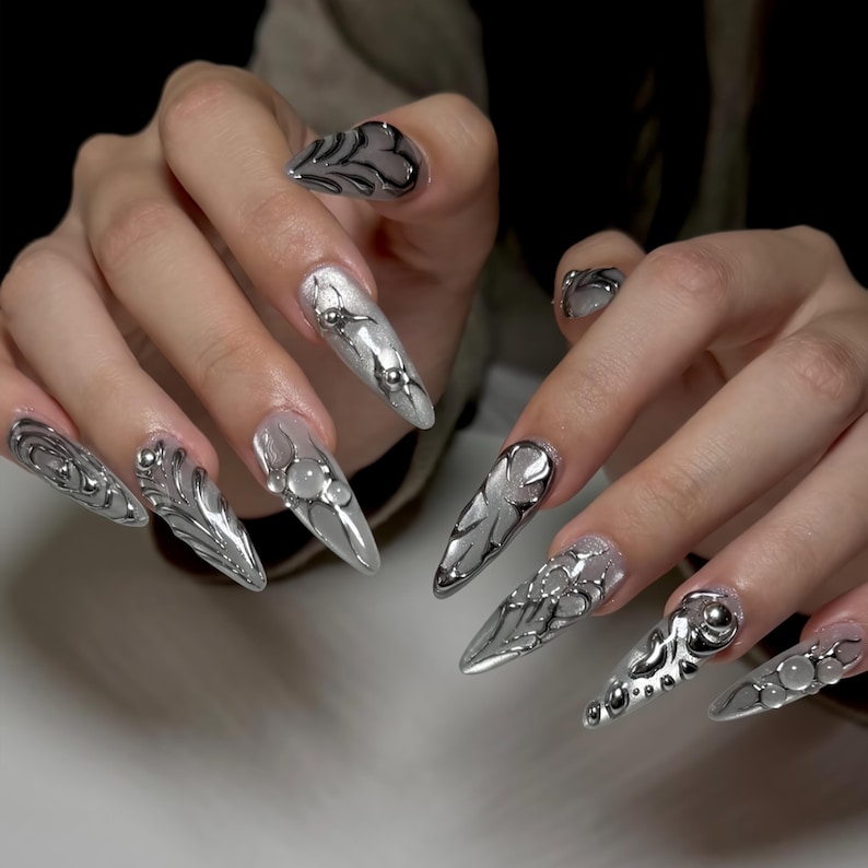 May include: Long, pointed silver and clear acrylic nails with intricate metallic designs. The nails feature a variety of embellishments, including small silver beads and swirling patterns. The overall aesthetic is futuristic and edgy.