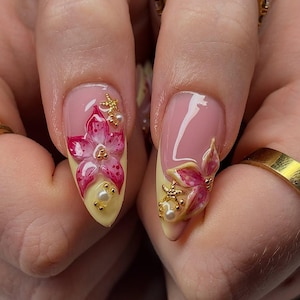 May include: Close-up of two manicured nails with a floral design. The nails are painted with a light pink base and a yellow tip. Each nail features a 3D pink flower with gold accents and small pearls.