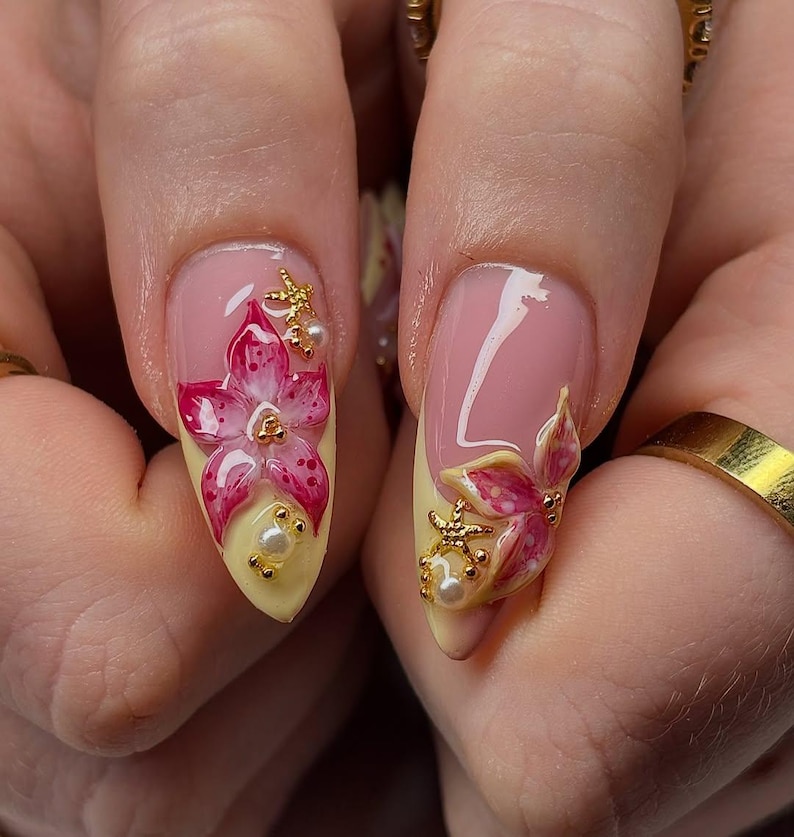 May include: Close-up of two manicured nails with a floral design. The nails are painted with a light pink base and a yellow tip. Each nail features a 3D pink flower with gold accents and small pearls.