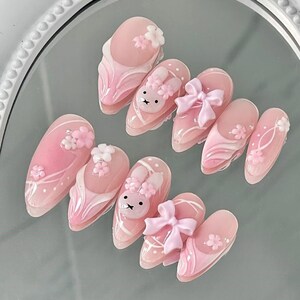 May include: A set of ten pink artificial nails with various designs. Some nails feature 3D white flowers, while others have a bunny face or a pink bow. The nails have a glossy finish and are arranged on a reflective surface.