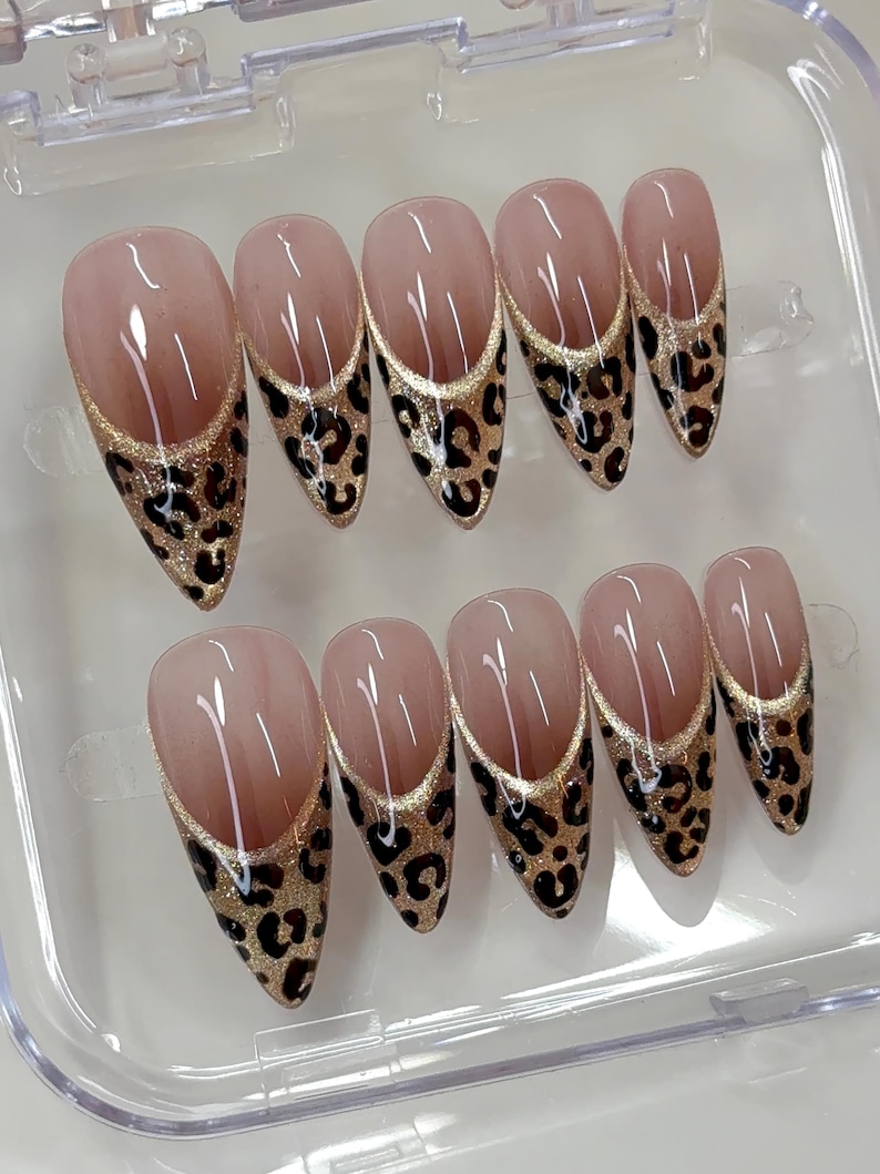 Puede incluir: Un juego de diez u&ntilde;as postizas en forma de almendra con una base de color nude. Cada u&ntilde;a presenta un dise&ntilde;o de punta francesa en forma de V profunda con un borde de purpurina dorada y un estampado de leopardo negro y marr&oacute;n en la punta.