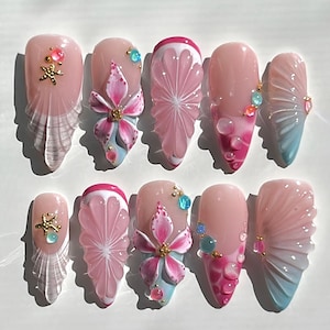 May include: A set of ten almond-shaped artificial nails. The nails feature a variety of designs, including pink and white citrus slices, pink flowers, and clear, fan-shaped accents. Some nails have gold starfish and jewel embellishments.