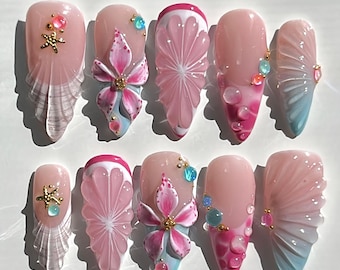 May include: A set of ten almond-shaped artificial nails. The nails feature a variety of designs, including pink and white citrus slices, pink flowers, and clear, fan-shaped accents. Some nails have gold starfish and jewel embellishments.