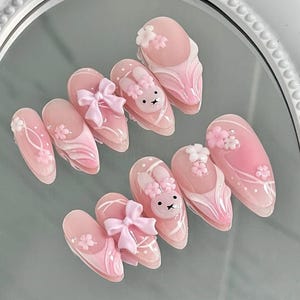 May include: A set of ten almond-shaped, press-on nails. Each nail is painted in shades of pink and white, with decorative elements including bows, bunny faces, and floral accents. The nails are arranged on a reflective surface.