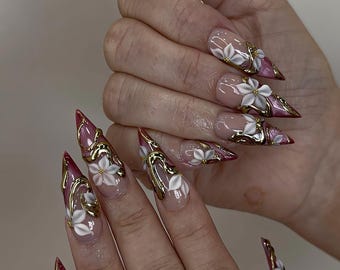 May include: Long, pointed fingernails with a clear base, adorned with white 3D flowers, gold accents, and burgundy tips. The nails feature intricate designs, creating a glamorous and artistic look. The nails are a form of nail art.