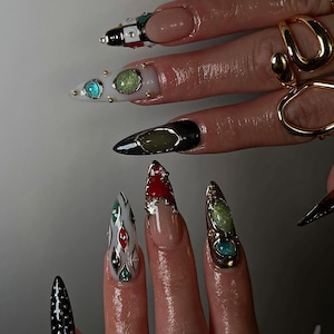 Könnte beinhalten: Nahaufnahme einer Hand mit langen, aufwendig verzierten Fingernägeln. Die Nägel weisen verschiedene Designs auf, darunter schwarze, weiße, rote und grüne Akzente sowie goldene und silberne Verzierungen. Einige Nägel haben edelsteinartige Details.