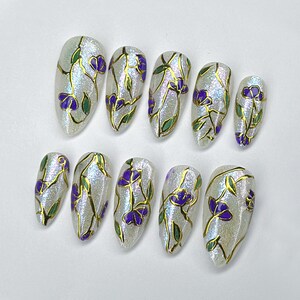 May include: Set of ten artificial nails with a shimmering, pearlescent finish. Each nail is adorned with a gold outline design featuring purple flowers and green leaves. The nails are oval-shaped.