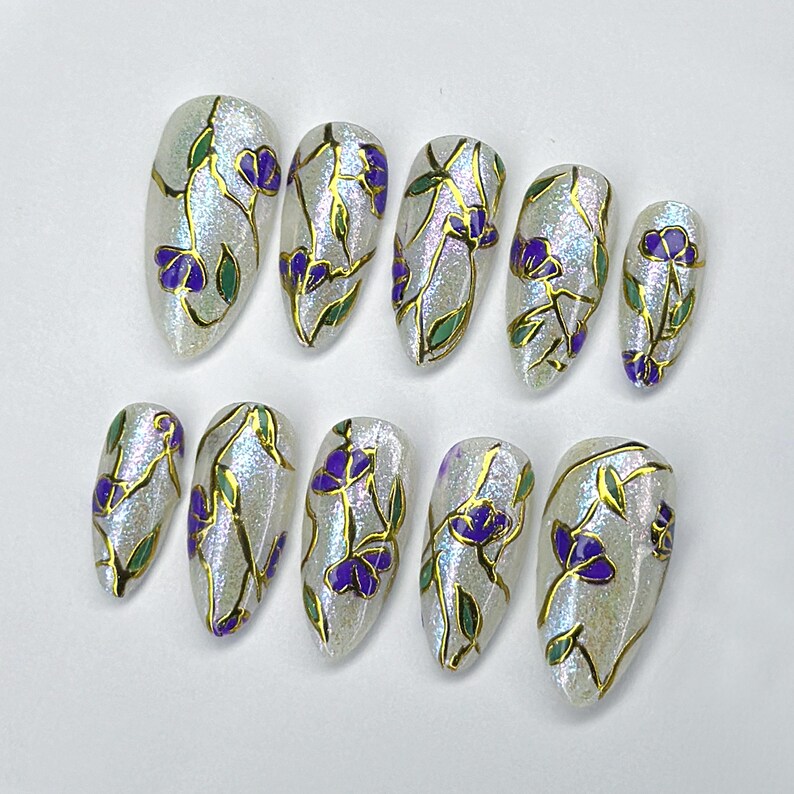 May include: Set of ten artificial nails with a shimmering, pearlescent finish. Each nail is adorned with a gold outline design featuring purple flowers and green leaves. The nails are oval-shaped.