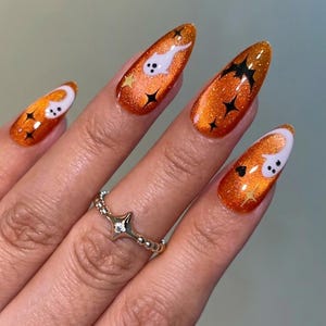 May include: Almond-shaped nails featuring a vibrant orange glitter polish base. The design includes white ghost figures, black bat silhouettes, and small black and gold star accents. A silver-toned ring with a star and moon motif is worn.