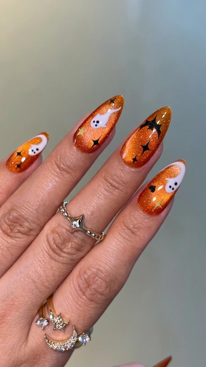 May include: Almond-shaped nails featuring a vibrant orange glitter polish base. The design includes white ghost figures, black bat silhouettes, and small black and gold star accents. A silver-toned ring with a star and moon motif is worn.