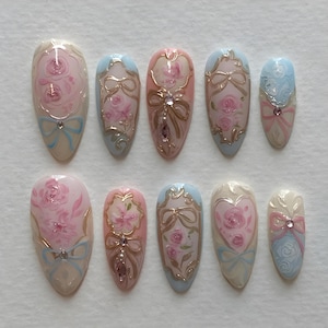 May include: A set of ten almond-shaped false nails. Each nail features a hand-painted design with pink roses, gold accents, and light blue bows. Some nails have a central jewel. The overall design is delicate and feminine.