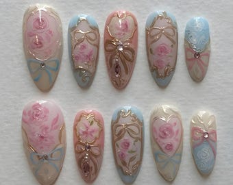 May include: A set of ten almond-shaped false nails. Each nail features a hand-painted design with pink roses, gold accents, and light blue bows. Some nails have a central jewel. The overall design is delicate and feminine.