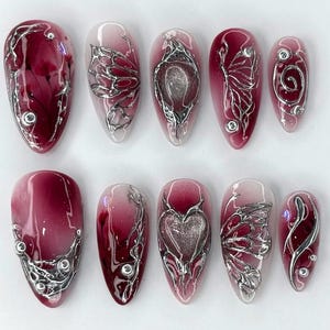 Puede incluir: Un conjunto de diez u&ntilde;as artificiales con un dise&ntilde;o degradado burdeos y transparente. Cada u&ntilde;a presenta adornos met&aacute;licos plateados, incluyendo espirales, alas de mariposa y formas de coraz&oacute;n. Las u&ntilde;as son de forma ovalada y tienen un acabado brillante.