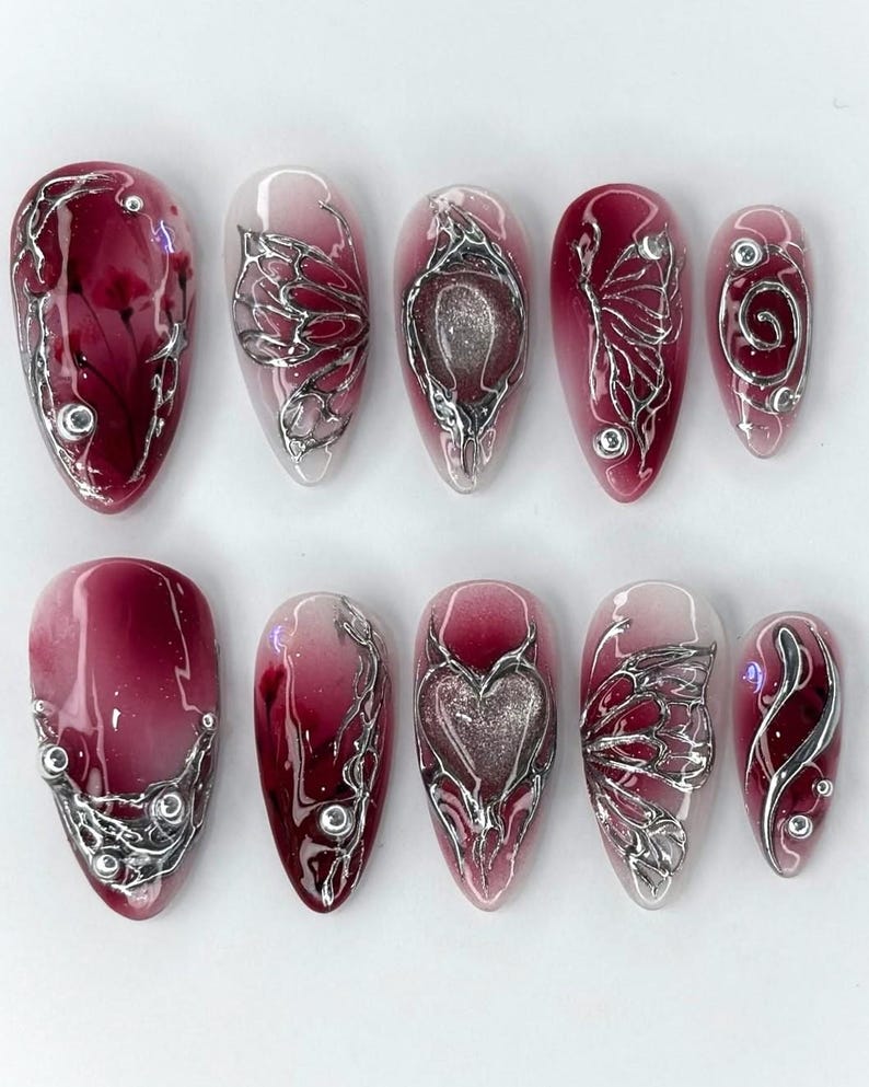 Puede incluir: Un conjunto de diez u&ntilde;as artificiales con un dise&ntilde;o degradado burdeos y transparente. Cada u&ntilde;a presenta adornos met&aacute;licos plateados, incluyendo espirales, alas de mariposa y formas de coraz&oacute;n. Las u&ntilde;as son de forma ovalada y tienen un acabado brillante.