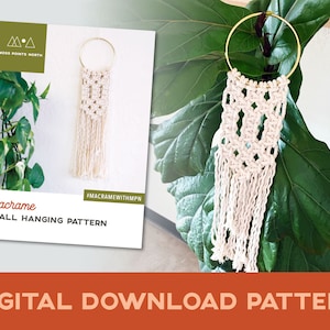 May include: A white macrame wall hanging with a geometric pattern, hanging from a gold hoop. The macrame is made with natural cotton cord and has a fringe at the bottom. The hanging is displayed against a white wall with a green plant in the background. The text "Macrame Wall Hanging Pattern" and "#macramewithmpn" are visible on the image.