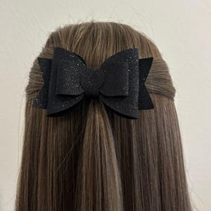 Op de afbeelding: Een zwarte glitter haar strik met een grote centrale lus en twee kleinere zijlussen. De strik is gebonden in het bruine haar van een persoon.