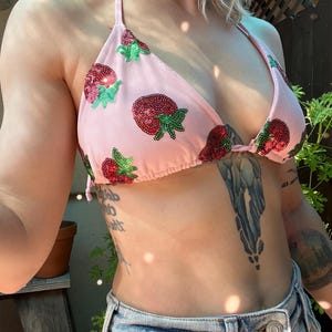 May include: Pink halter bikini top adorned with sequined strawberry designs. The strawberries feature red sequins and green leaf accents. The bikini top is paired with denim shorts.