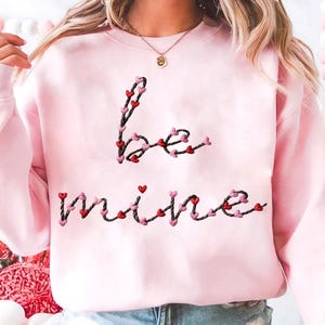 May include: A light pink sweatshirt with the embroidered words "be mine" in a cursive font. The letters are decorated with small red and pink hearts, creating a romantic and festive design. The sweatshirt is a casual, comfortable garment.
