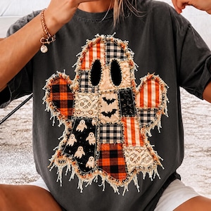 May include: A charcoal gray short-sleeve t-shirt featuring a large, central ghost-shaped graphic. The ghost is constructed from a patchwork of various fabrics, including orange and black plaid, striped patterns, lace, and black fabric with bats.