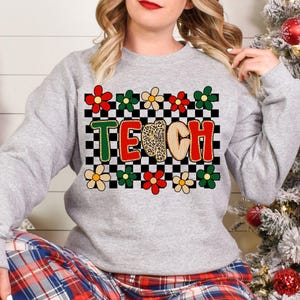 Pu&ograve; includere: Una felpa grigia con la scritta "TEACH" in un design colorato. Le lettere sono bordate di verde, rosso e beige, con una "C" con stampa leopardata. Il design include uno sfondo a scacchi e accenti floreali in rosso, verde e crema.
