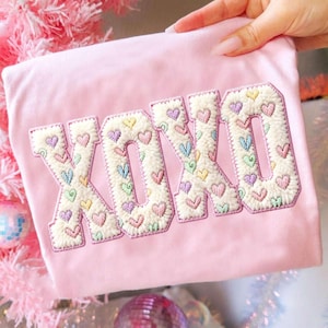 May include: A light pink, plush fabric item with the embroidered letters "XOXO". The letters are filled with a fluffy, off-white material and decorated with small, colorful embroidered hearts. The item is held by a hand.