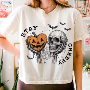 May include: Cream-colored t-shirt with a Halloween-themed graphic. The design features a pumpkin-shaped jack-o'-lantern, a skull, bats, and the words "STAY CREEPY". The artwork is in black and orange.