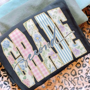 May include: A dark grey t-shirt featuring the words "Second Grade" in large, decorative letters. The letters are filled with floral patterns, pink gingham, and blue stripes. The word "second" is in a cursive font.