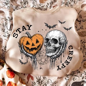 May include: Beige sweatshirt with a Halloween-themed graphic. The design features a heart-shaped jack-o'-lantern held by skeletal hands, a skull, bats, and a spiderweb. The words "STAY CREEPY" are printed on the sweatshirt.