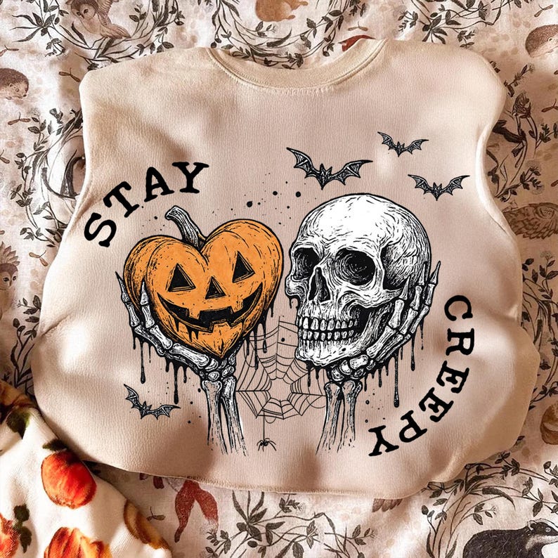 May include: Beige sweatshirt with a Halloween-themed graphic. The design features a heart-shaped jack-o'-lantern held by skeletal hands, a skull, bats, and a spiderweb. The words "STAY CREEPY" are printed on the sweatshirt.