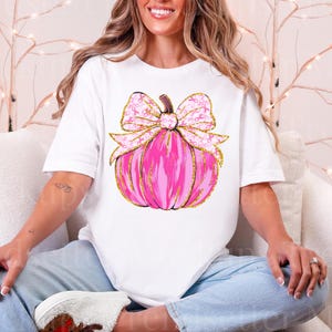 May include: White t-shirt featuring a pink pumpkin design with a gold outline and a bow adorned with pink ribbons. The pumpkin design is centered on the shirt, creating a visually appealing graphic.