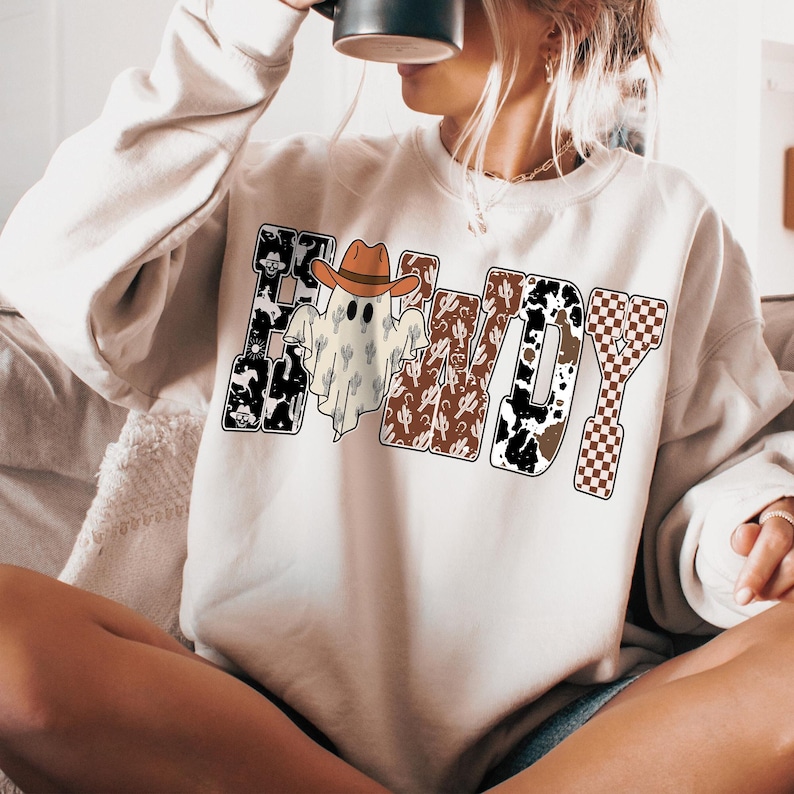 K&ouml;nnte beinhalten: Cremefarbenes Sweatshirt mit dem Wort "HOWDY" in einem Western-Design. Das "H" ist schwarz mit Totenkopf-Akzenten, das "O" zeigt einen Geist mit Cowboyhut und das "W" hat ein Kaktusmuster.