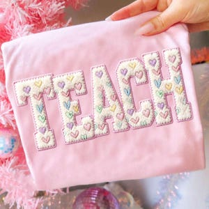 May include: A light pink t-shirt with the word "TEACH" embroidered in a white, textured font. Each letter is filled with colorful heart shapes. The shirt is held up by a hand, with a pink Christmas tree in the background.