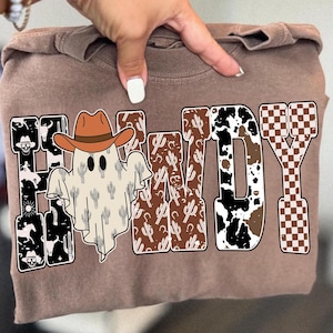 May include: A brown sweatshirt with the word "Howdy" in a Western-themed design. The "H" and "Y" are black and white, the "O" and "W" are brown with cactus patterns, and the "D" is cow print. A ghost wearing a cowboy hat is in the center.