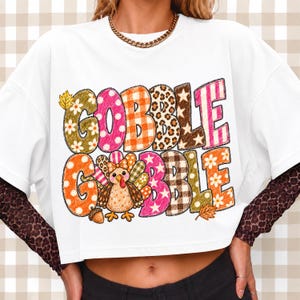 May include: A white short-sleeved t-shirt featuring the text GOBBLE GOBBLE in large, multi-patterned letters including floral, polka dot, plaid, leopard print, and star designs. A cartoon turkey with a bow is integrated into the graphic.