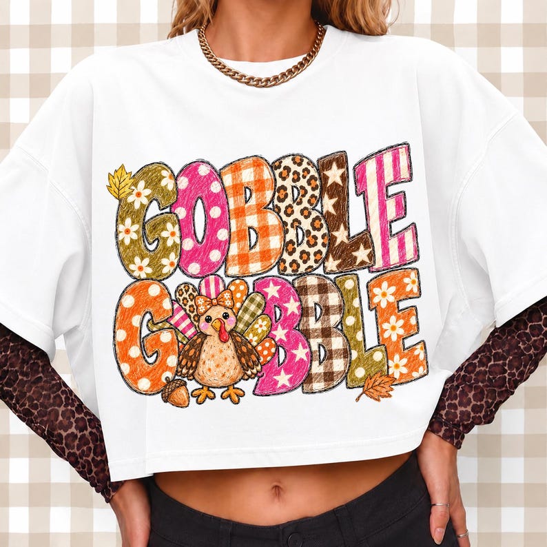 May include: A white short-sleeved t-shirt featuring the text GOBBLE GOBBLE in large, multi-patterned letters including floral, polka dot, plaid, leopard print, and star designs. A cartoon turkey with a bow is integrated into the graphic.