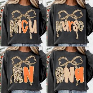 May include: Dark gray sweatshirts with text and bow designs. The sweatshirts feature the words "NICU", "Nurse", "RN", and "CNA" in orange and leopard print lettering. A leopard print bow is above each word.