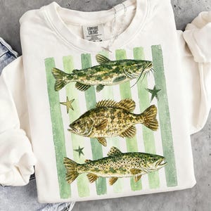 May include: Cream-colored sweatshirt featuring a watercolor design of three fish: a catfish, a bass, and a trout, set against green vertical stripes and star accents. The sweatshirt has a crew neck and long sleeves. The brand name "Comfort Colors" is visible on the tag.
