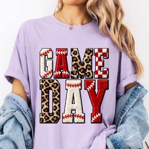 May include: A light purple t-shirt with the words "GAME DAY" in a baseball-themed design. The letters are a mix of leopard print, red, white, and baseball stitching. The shirt has short sleeves and a relaxed fit.