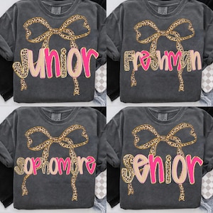 May include: Four dark gray t-shirts with different grade level designs. Each shirt features a leopard print bow above a word in pink and tan: "Junior", "Freshman", "Sophomore", and "Senior".