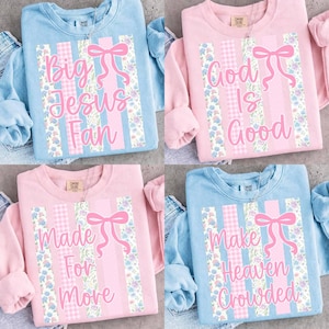 Puede incluir: Cuatro sudaderas de colores pastel con texto inspirador. Cada sudadera presenta un diseño central con un lazo y rayas verticales. El texto en las camisas dice: "Big Jesus Fan", "God Is Good", "Made For More" y "Make Heaven Crowded".