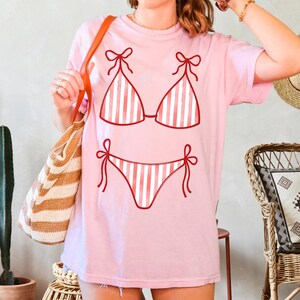 May include: A pale pink t-shirt with a red and white striped bikini design. The bikini top and bottom are outlined in red with bow details. The shirt is a casual, comfortable style.