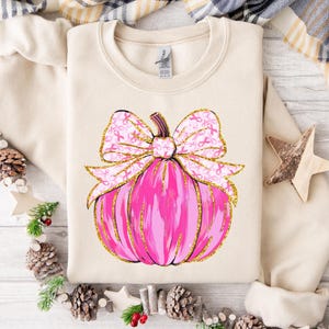 May include: Cream-colored sweatshirt featuring a pink pumpkin design with a gold glitter outline and a bow adorned with pink ribbons. The pumpkin is a symbol of autumn and the ribbons represent breast cancer awareness.