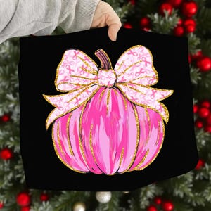 May include: A pink pumpkin design on a black background. The pumpkin has a gold glitter outline and a bow with pink ribbons. The bow is decorated with pink ribbons. The pumpkin is a symbol of breast cancer awareness.