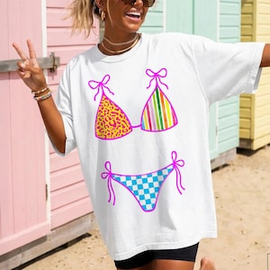 以下が含まれることがあります： カラフルなビキニのグラフィックが特徴の白いTシャツ。ビキニトップはヒョウ柄のデザインと縦縞模様。ビキニボトムは青と白のチェック柄。グラフィックはピンクで縁取られています。