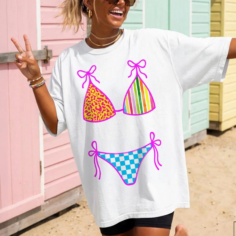 以下が含まれることがあります： カラフルなビキニのグラフィックが特徴の白いTシャツ。ビキニトップはヒョウ柄のデザインと縦縞模様。ビキニボトムは青と白のチェック柄。グラフィックはピンクで縁取られています。